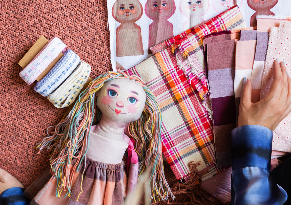 Zdjęcie lalki Astrid leżącej wśród kolorowych materiałów i wstążek przygotowanych do planowania kolekcji.
